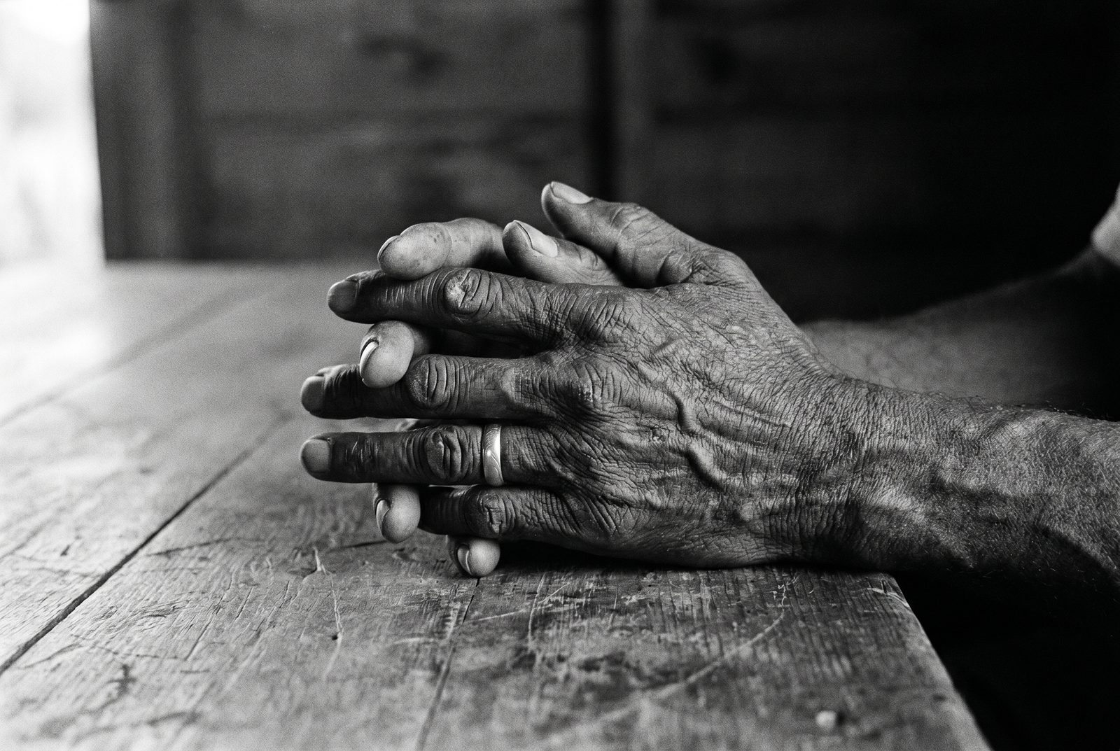 Black-and-white close-up of the weathered hands of an elderly Black man clasped on a wooden table.